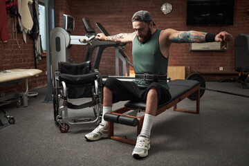 Man wearing green tank top and fitness belt exercising on gym equipment with his wheelchair nearby....