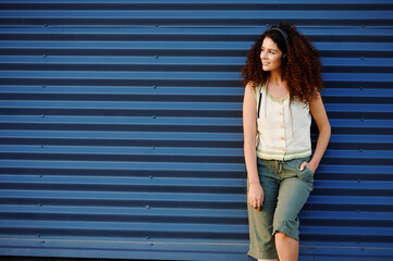 confident woman wearing headphones looks to side against blue metal wall backdrop, exuding relaxed...