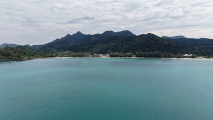 Turquoise water gently laps against the sandy shore of a tropical island, with lush green mountains rising majestically in the background under a cloudy sky, creating a serene and picturesque scene