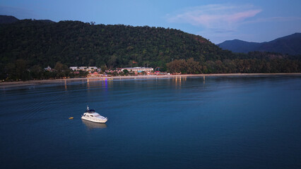 Motor yacht floating near a tropical beach at dusk in thailand, with lights reflecting on the calm water and forested hills in the background
