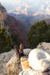 A tourist is sitting at the edge of the Grand Canyon, enjoying a meal. The camera’s focus is set...
