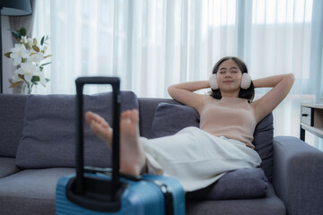 Young woman wearing casual clothes is lying on a comfortable sofa with her feet resting on her suitcase, listening to music with headphones and dreaming of her next trip