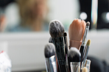 Close-up of various makeup brushes in soft focus with a blurred background of a mirror and a person, creating a professional beauty workspace atmosphere.