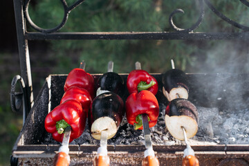 Obraz premium Close-up of red bell peppers and eggplants grilling on skewers over hot coals in an outdoor metal barbecue, with smoke rising in natural daylight.