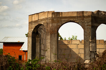 Weathered, unfinished concrete building with arched windows stands overgrown with vegetation, with...