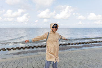 Smiling happy woman walking by Nord sea coast wear beige coat  sunglasses enjoys windy day cloudy sky background. Female person have fun hiking adventure travel at Baltic sea seaside