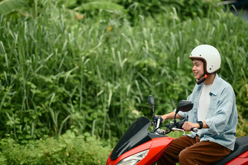 Smiling young man in helmet riding a red motorbike along a rural road