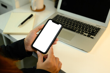 Close up of hands holding smartphone with blank white screen next to laptop
