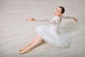 Young female ballerina with brown hair in white tutu, seated elegantly on wooden floor. Soft lighting enhances her poised expression and delicate pose, casting gentle shadows © TRAVELARIUM