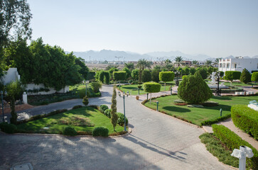 Fototapeta premium Spacious resort garden offers lush greenery and winding paths, surrounded by towering mountains in distance. Warm daylight enhances natural beauty with verdant tones