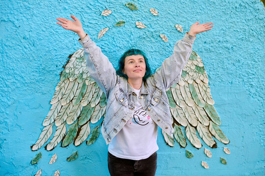 Fototapeta Young woman with dyed hair poses joyfully in front of blue mural. Angel wings painted on wall create illusion of wings on her back. Denim jacket enhances casual look