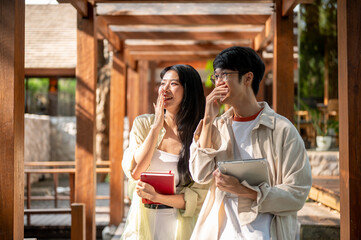 Fototapeta premium Smiling asian woman and a male friend standing with a hand covered mouth in garden wooden corridor.