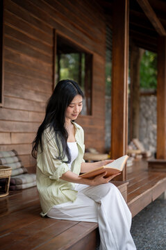 Fototapeta Long hair asian woman sitting cross lagged while reading a book at wooden house's terrace.