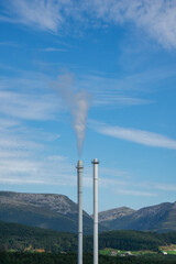 Two industrial chimneys release smoke into the clear blue sky with the lush forest and rocky mountains of the Norwegian landscape in background