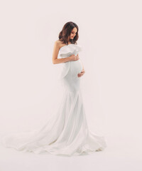 Happy pregnant woman in white dress gently hugs touch her belly with hands. Light beige studio background. Beautiful pregnancy, motherhood, expecting a baby, family health concept. Free space for text