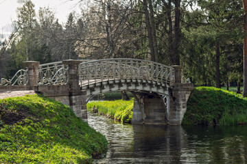 Elegant wrought iron bridge spans calm park canal, framed by greenery and old trees, offering serene atmosphere in spring sunlight.