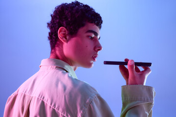 Rear view photo. Teen boy holding phone near mouth as if using voice assistant in colorful neon...