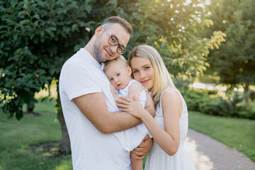 Fototapeta premium Portrait of a happy young family spending time together on nature, on vacation, outdoors. Mom, dad holds daughter stand on the grass. The concept of family holiday.