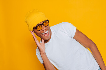 Excited young black african american man with yellow beanie and glasses holding hand to ear,...