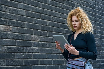 Young woman with curly blonde hair using digital tablet against modern black brick wall background, standing outdoors, focused and stylish, casual urban lifestyle and technology usage concept.