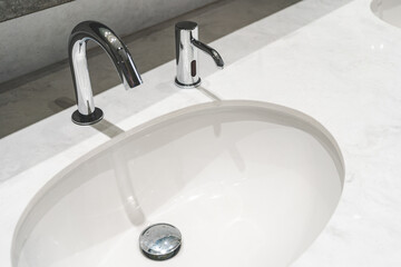 Modern Chrome Automatic Faucet and Soap Dispenser in Clean Public Restroom. Chrome water tap on white ceramic wash basin.