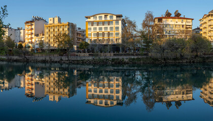 Evening view of Manavgat river with mirror-like reflection of buildings and trees along the promenade. Warm sunset light creates peaceful atmosphere. Manavgat, Turkey.