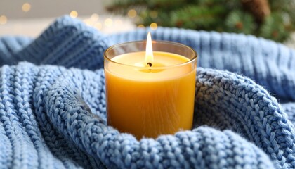 A lit yellow candle in a glass holder sits on a cozy blue knitted blanket, with soft festive lights and greenery in the background.