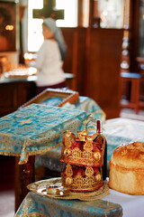 Ornate orthodox wedding golden crowns, bread, and wine on embroidered cloth in Orthodox church interior with blurred praying woman in background.