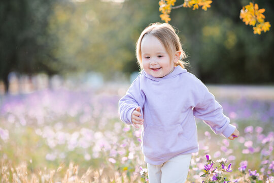 Smiling cute kid girl 3-4 year old wearing hoodie running in meadow over flowers outdoors. Summer season. Childhood.
