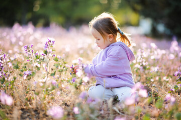 Smiling cute kid girl 3-4 year old wearing hoodie playing in field with flowers outdoors. Summer season. Childhood.
