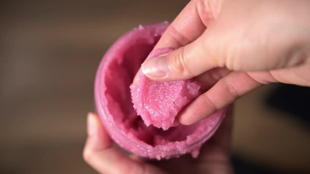 Female hand scoops pink raspberry sugar body scrub from jar showing its texture. Smooth, creamy texture ideal for exfoliating and nourishing skin. Body scrub for skincare, wellness and spa concepts.