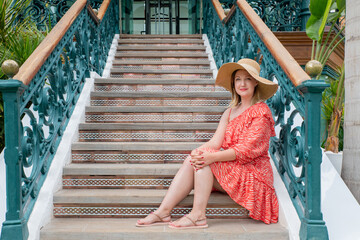 Stylish woman in a red patterned dress and a wide-brim straw hat while sitting comfortably on...