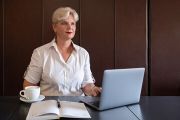 Mature woman with short blond hair engaging in digital work from a quiet stylish workplace with a hot coffee aside, a sleek laptop and a planner with pen, concept for business coaching or freelancing