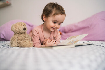 A happy toddler lies on a comfortable bed with a teddy bear, engrossed in reading a colorful picture book. This young child is enjoying quiet time and learning.