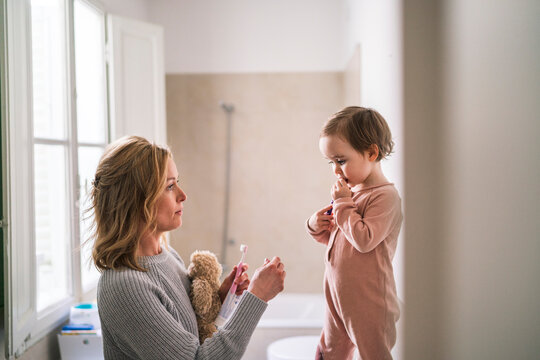 A mother and her toddler child are in the bathroom. The mother holds a toothbrush and toothpaste while the child holds their own toothbrush, preparing to brush their teeth.