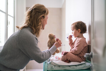 Mother and toddler child brush teeth together in the bathroom. The woman holds a teddy bear with a toothbrush, teaching the child good dental hygiene habits.