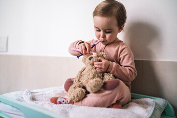 A toddler sits and brushes a teddy bear's nose with a toothbrush. The child holds another toothbrush near their mouth, practicing dental hygiene skills.