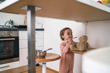 A young child in a pink outfit stands at a table, feeding a teddy bear with a spoon. The child is engaged in imaginative play in a bright kitchen setting.