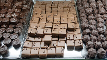 Display in a chocolatery or a coffee shop of a large assortment of in-house crafted chocolates, gourmet selection of dark rounds with a swirl on top, smooth milk squares with cacao nibs and nuts balls