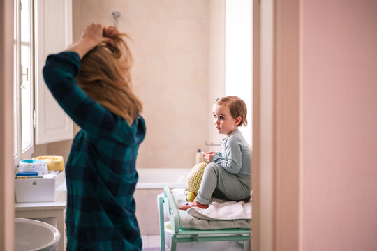 A mother and her young daughter spend time together in the bathroom during their morning routine. The mother fixes her hair while the child sits nearby.