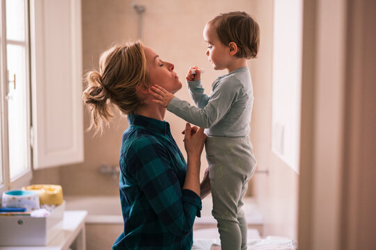 A mother and her toddler child share a sweet moment together indoors. The child stands and holds a small object, while the mother supports them gently.