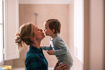 A loving mother holds her young child who leans in to give her a kiss. This intimate moment between parent and toddler shows a strong family connection and affection indoors.