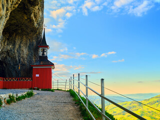 Beautiful red chapel nestled under a rocky cliffside near a scenic overlook with lush green hills and a colorful sky