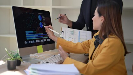 A woman is sitting at a desk with a computer monitor and a keyboard. She is looking at a graph on the monitor - Powered by Adobe