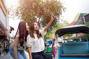 Two Young Asian Women Taking a Fun Selfie Together on City Street with Colorful Rickshaws Background