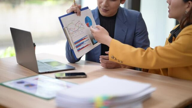 A man is presenting a report to a woman, showing her a piece of paper with various graphs and charts on it. The woman is attentively looking at the paper