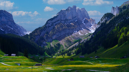 Majestic mountains rise above a lush valley with scattered farmhouses in a picturesque landscape under blue skies