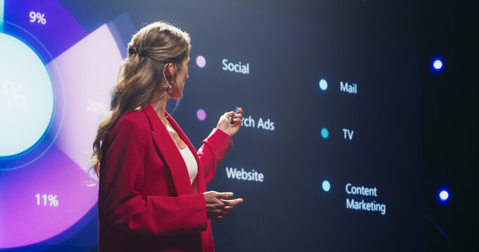 Beautiful Adult Female in a Red Suit Making a Passionate Lecture with a Big Screen Showing Different Analytical Data in the Background. Representative Sharing a Company Earning Report to Shareholders