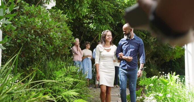 Guests watching bride walking under pergola, man escorting and officiant handing ceremony binder