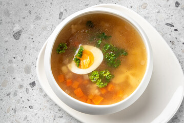 A bowl of clear vegetable soup with diced carrots, potatoes, parsley, and half a boiled egg, served on a white plate against a neutral speckled gray background.